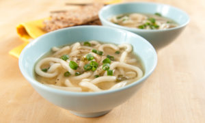 Miso soup with noodles in blue bowl sitting on light wooden table.