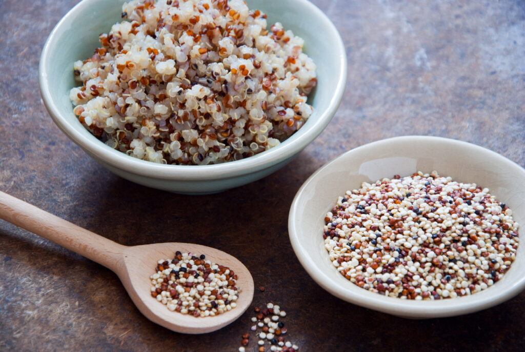 Bowls of multi-coloured Quinoa seeds (one cooked and one raw) on table next to wooden spoon containing raw quinoa seeds.