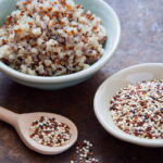 Bowls of multi-coloured Quinoa seeds (one cooked and one raw) on table next to wooden spoon containing raw quinoa seeds.